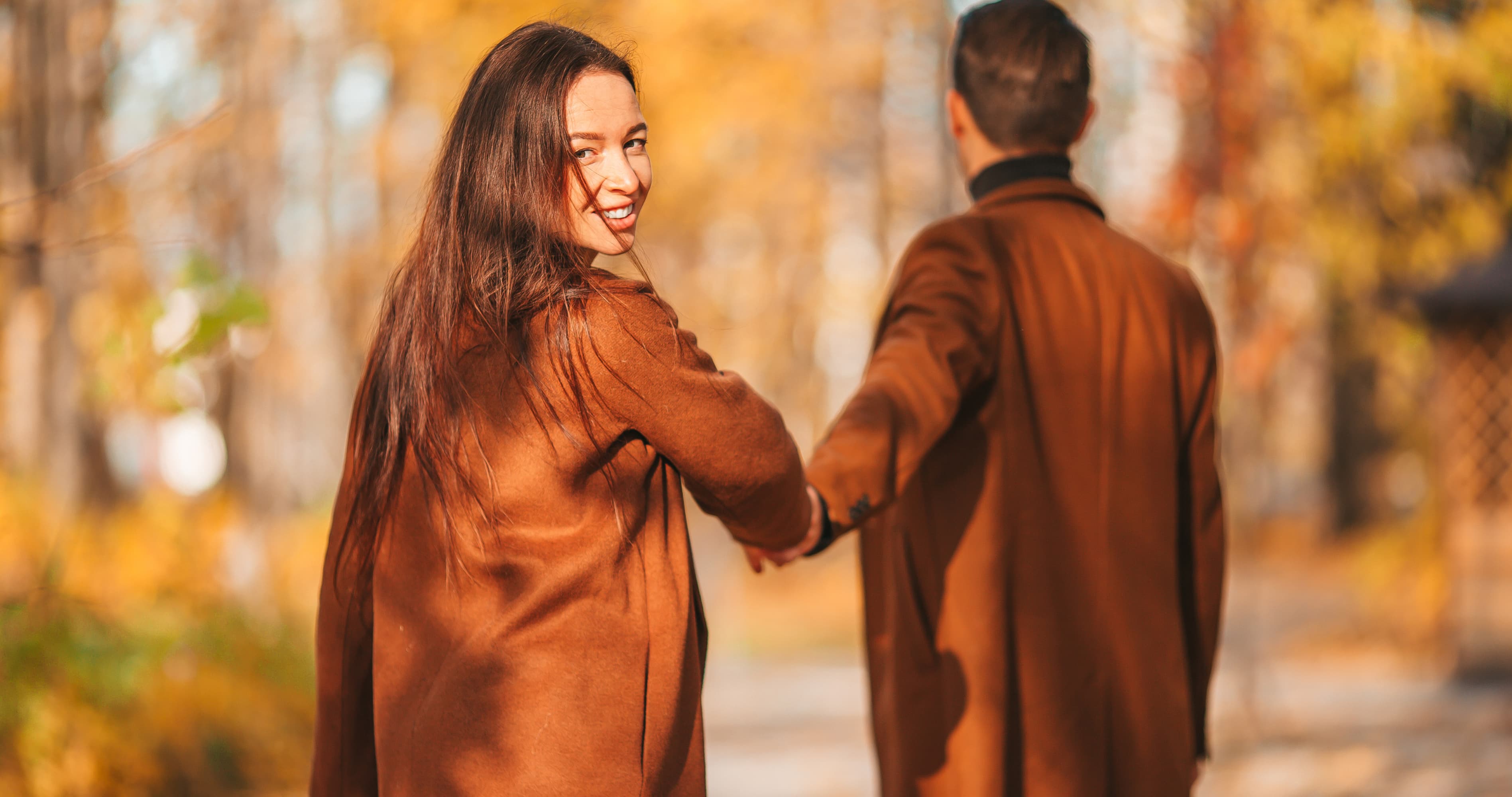 A happy couple holding hands and walking together through a beautiful autumn park, embodying how to feel secure in a relationship as the woman smiles back.