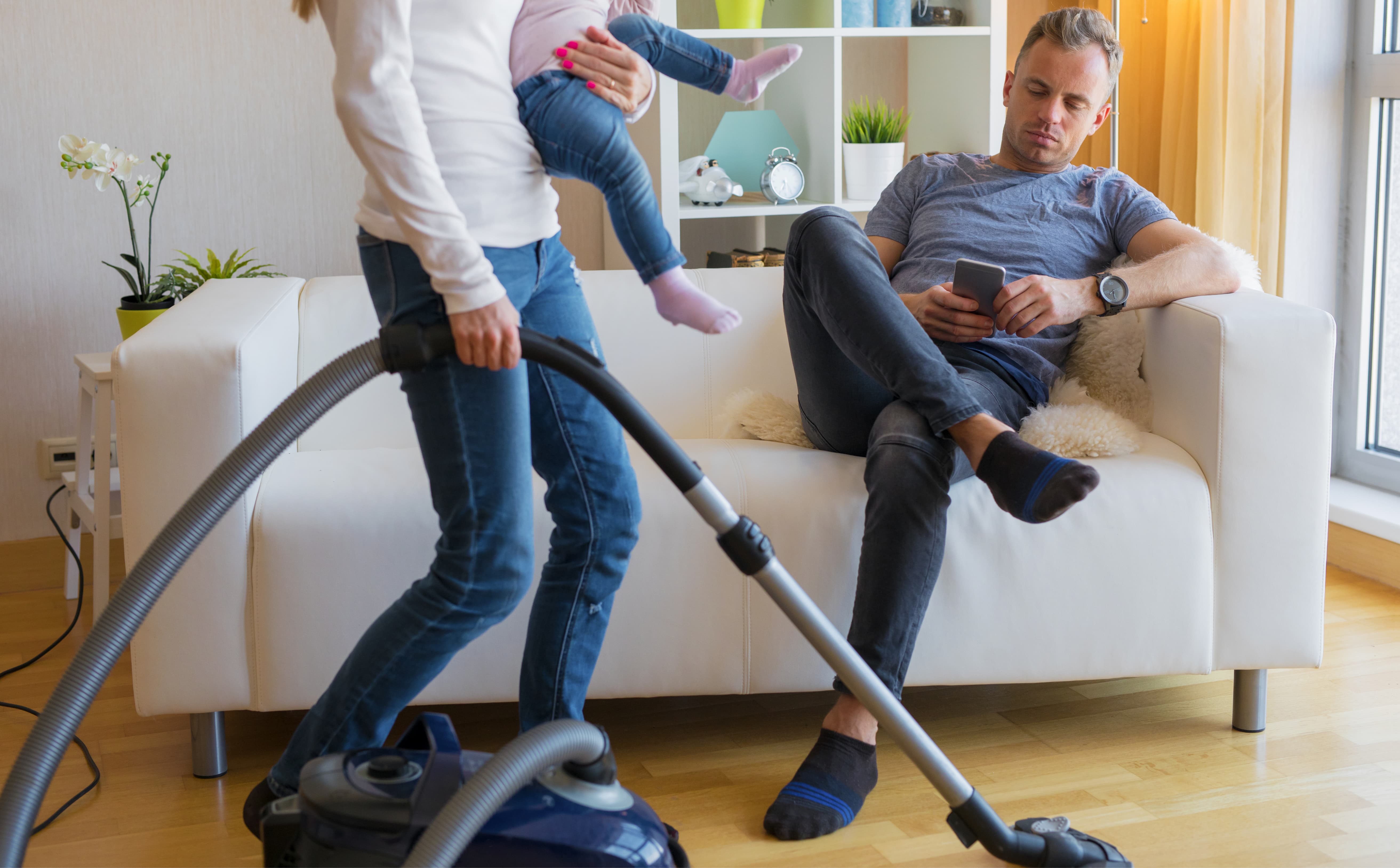 Woman vacuuming while carrying a child as her husband relaxes on the couch, showing an unfair split in marriage and unequal household responsibilities.
